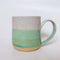 A handmade ceramic mug with a colorblock design featuring a white top and a speckled green middle section, displayed on a light gray background. The mug has a large, rounded handle, a speckled texture, and is made in Mississippi Mills, Ontario, Canada.