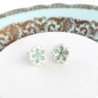 A pair of tiny, flower-shaped stud earrings with delicate mint green petals outlined in white and a touch of 22K gold detailing in the center. They are displayed on a white surface with a decorative mint green and gold plate partially visible in the background.