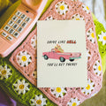 Notebook with 'Drive Like Hell, You'll Get There!' text on a pink phone and crocheted surface with flowers.