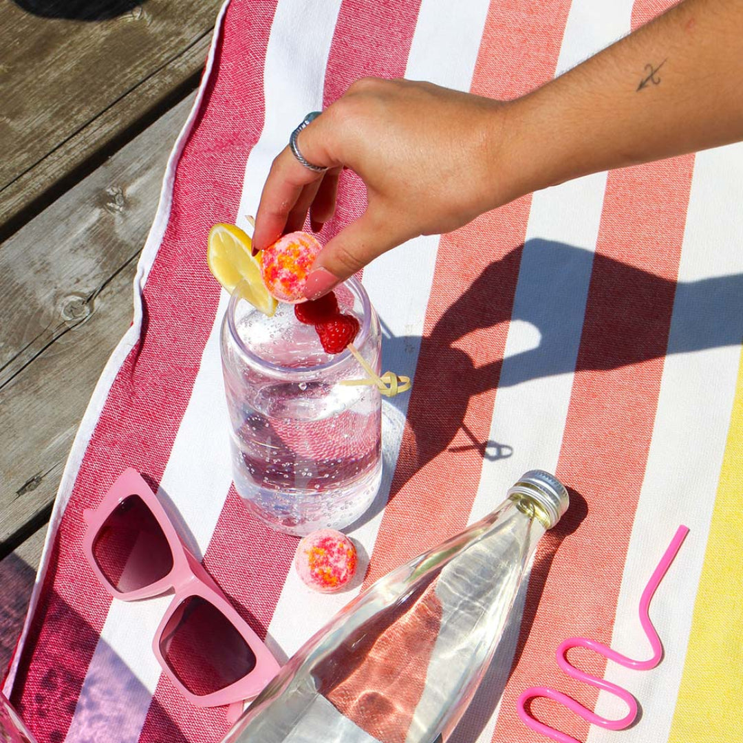 Hand placing a lemon wedge into a glass of water on a striped towel with sunglasses and a bottle.