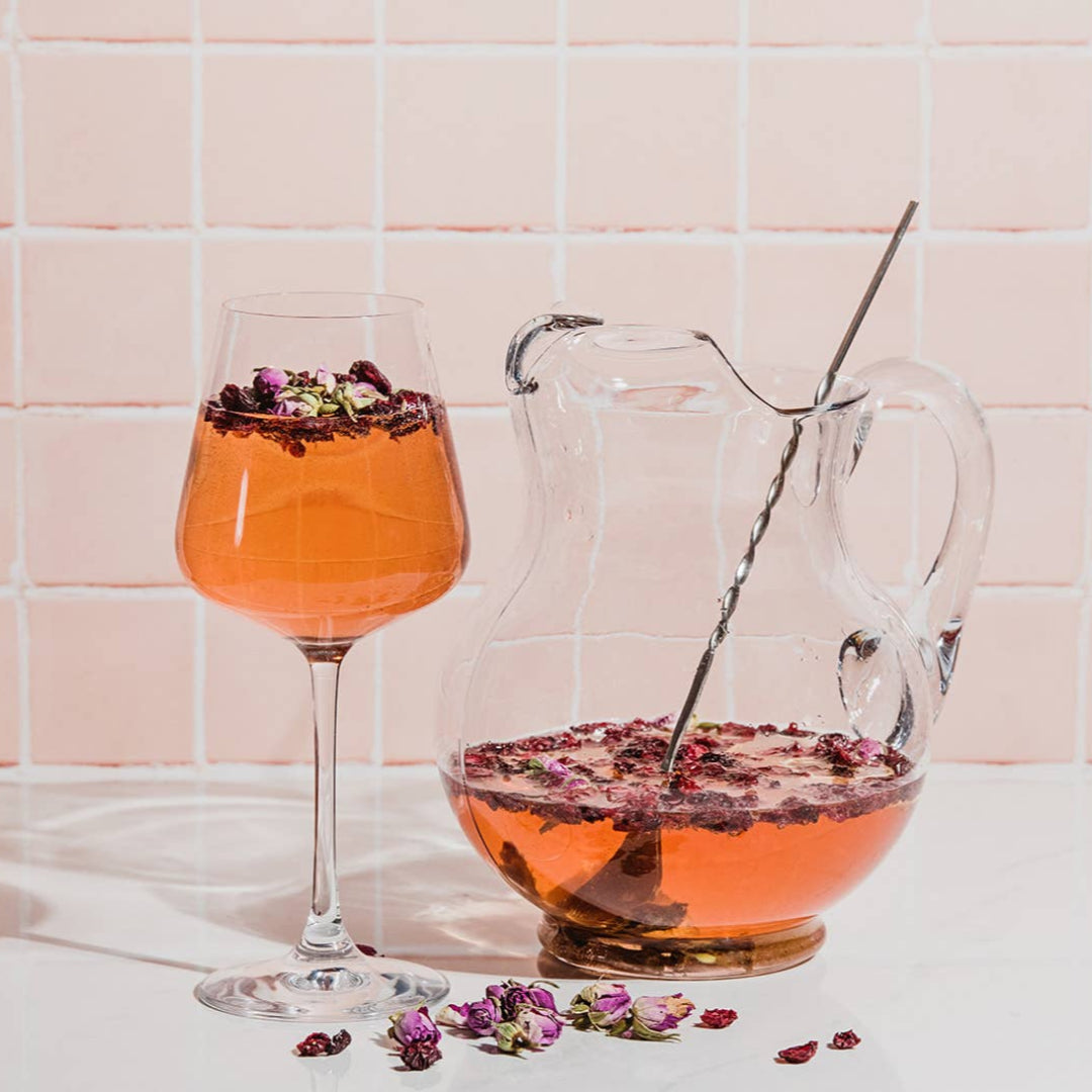 Glass of orange cocktail with floral garnish next to a pitcher of the same cocktail on a tiled wall background