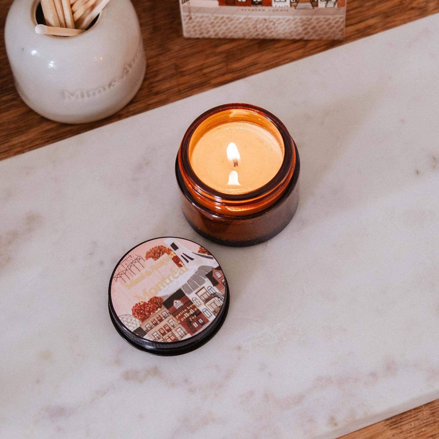 Candle with decorative lid on a marble surface, next to a container of matches and a small book.