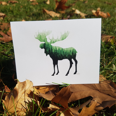 A white card with a black moose silhouette stands upright on green grass with fallen brown leaves. The moose's silhouette is filled with a vibrant green watercolor forest scene.