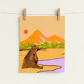 A vertical art print featuring a stylized illustration of a brown bear sitting by a lake with mountains and a sun in the background, hanging from two wooden clothespins on a white wall.
