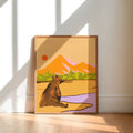 A framed art print featuring a stylized brown bear in a desert landscape with mountains, trees, and a purple foreground, leaning against a white wall on a light wood floor.