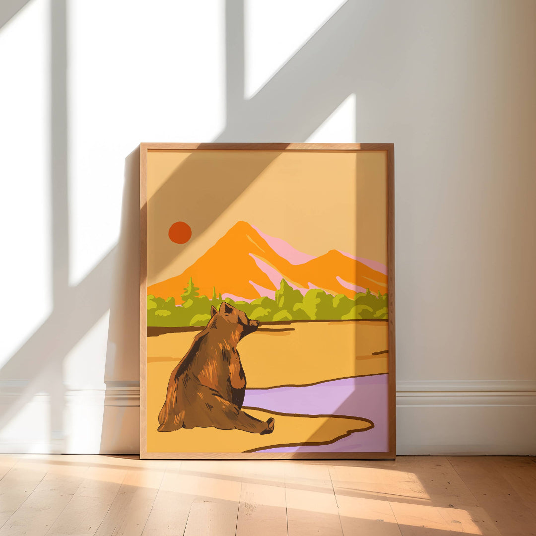 A framed art print featuring a stylized brown bear in a desert landscape with mountains, trees, and a purple foreground, leaning against a white wall on a light wood floor.