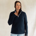 A woman wearing a navy blue quarter-zip pullover with white piping along the collar, standing against a neutral light beige fabric backdrop.