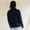 A woman viewed from behind wearing a dark navy long-sleeve quarter-zip fleece jacket and light blue jeans, standing against a neutral beige textured backdrop.