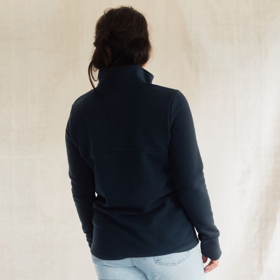 A woman viewed from behind wearing a dark navy long-sleeve quarter-zip fleece jacket and light blue jeans, standing against a neutral beige textured backdrop.