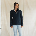 A woman modeling a dark navy quarter-zip fleece pullover with white contrast stitching and a white zipper trim, paired with light-wash blue jeans, standing against a draped beige fabric backdrop.
