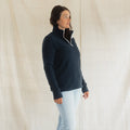 A woman modeling a navy blue quarter-zip fleece pullover with a white zipper trim, paired with light-wash jeans, standing against a draped off-white fabric backdrop.
