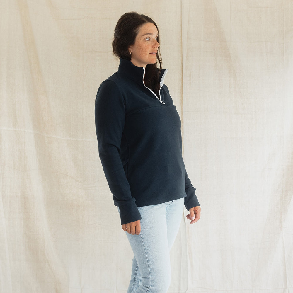 A woman modeling a navy blue quarter-zip fleece pullover with a white zipper trim, paired with light-wash jeans, standing against a draped off-white fabric backdrop.