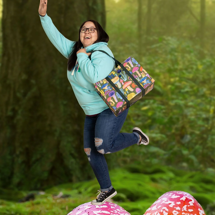 A large, reusable tote bag with a mushroom pattern. The bag is olive green with black handles. It is covered in a variety of colorful mushrooms in different shapes and sizes.