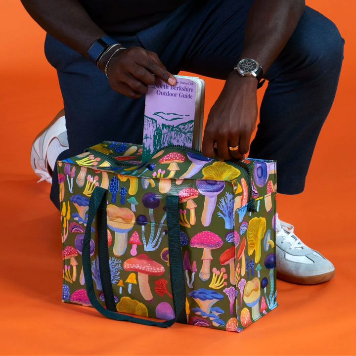 A large, reusable tote bag with a mushroom pattern. The bag is olive green with black handles. It is covered in a variety of colorful mushrooms in different shapes and sizes.