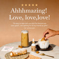 A styled coffee gift set on a marble table featuring an iced latte, a cup of hot coffee topped with whipped cream and a pastry, a pastry on a plate, a small dropper bottle, and a card, set against a warm beige background with promotional text.