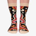 A pair of colourful women's crew socks with a black background featuring a retro-inspired celestial design with stars, moons, and rainbows in shades of orange, pink, yellow, and white. One sock displays a cartoon sun wearing sunglasses with rosy cheeks and the text "I'D RATHER BE NAPPING" in orange and white letters. The toe and cuff of the sock are a light beige.