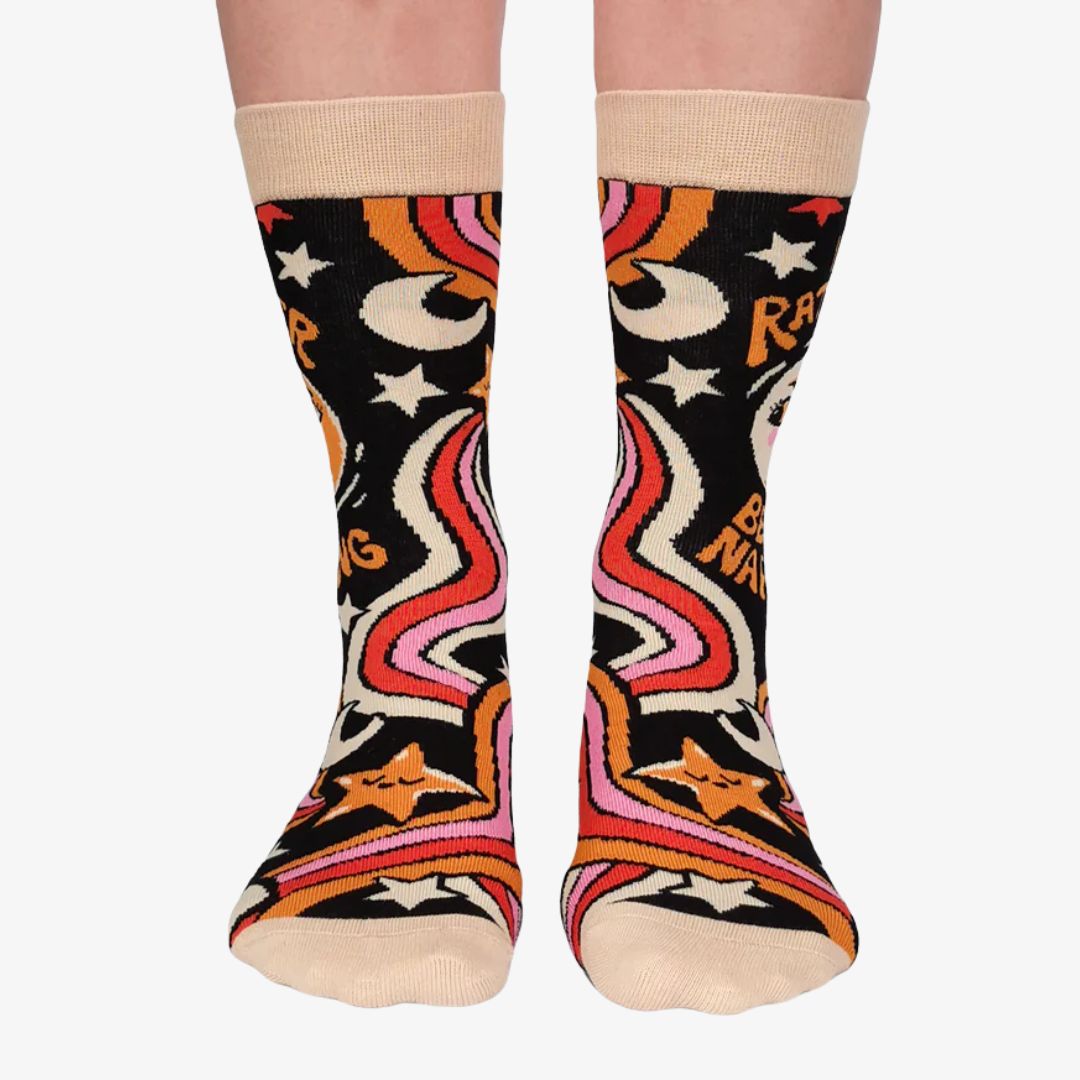 A pair of colourful women's crew socks with a black background featuring a retro-inspired celestial design with stars, moons, and rainbows in shades of orange, pink, yellow, and white. One sock displays a cartoon sun wearing sunglasses with rosy cheeks and the text "I'D RATHER BE NAPPING" in orange and white letters. The toe and cuff of the sock are a light beige.