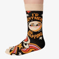 A pair of colourful women's crew socks with a black background featuring a retro-inspired celestial design with stars, moons, and rainbows in shades of orange, pink, yellow, and white. One sock displays a cartoon sun wearing sunglasses with rosy cheeks and the text "I'D RATHER BE NAPPING" in orange and white letters. The toe and cuff of the sock are a light beige.