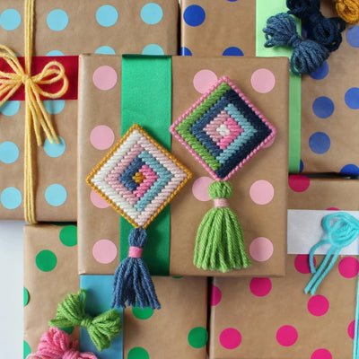 A flat lay showing several wrapped gifts with colorful polka dot wrapping paper. Two handmade cross-stitch ornaments, one pink and one blue, are placed on top of the gifts. Each ornament has a colorful tassel attached.