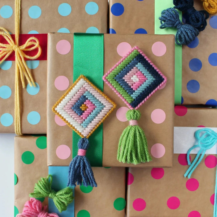A flat lay showing several wrapped gifts with colorful polka dot wrapping paper. Two handmade cross-stitch ornaments, one pink and one blue, are placed on top of the gifts. Each ornament has a colorful tassel attached.