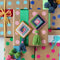 A flat lay showing several wrapped gifts with colorful polka dot wrapping paper. Two handmade cross-stitch ornaments, one pink and one blue, are placed on top of the gifts. Each ornament has a colorful tassel attached.