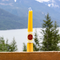 Two yellow beeswax candles standing upright on a wooden railing with a scenic mountain and lake background. The candles have a label that says 