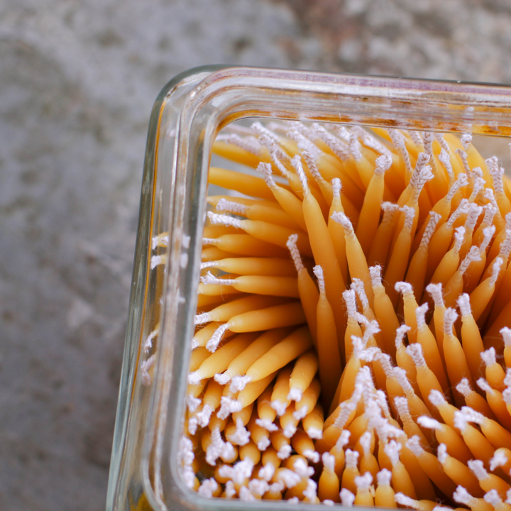A close-up of a glass jar filled with many thin, yellow beeswax candles. The candles are tightly packed together and their wicks are visible.