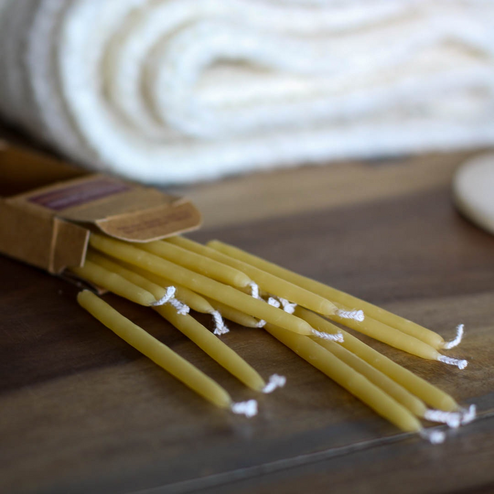 A cardboard box of birthday candles open on a wooden surface. The candles are yellow beeswax and are spilling out of the box. In the background, there is a white towel.