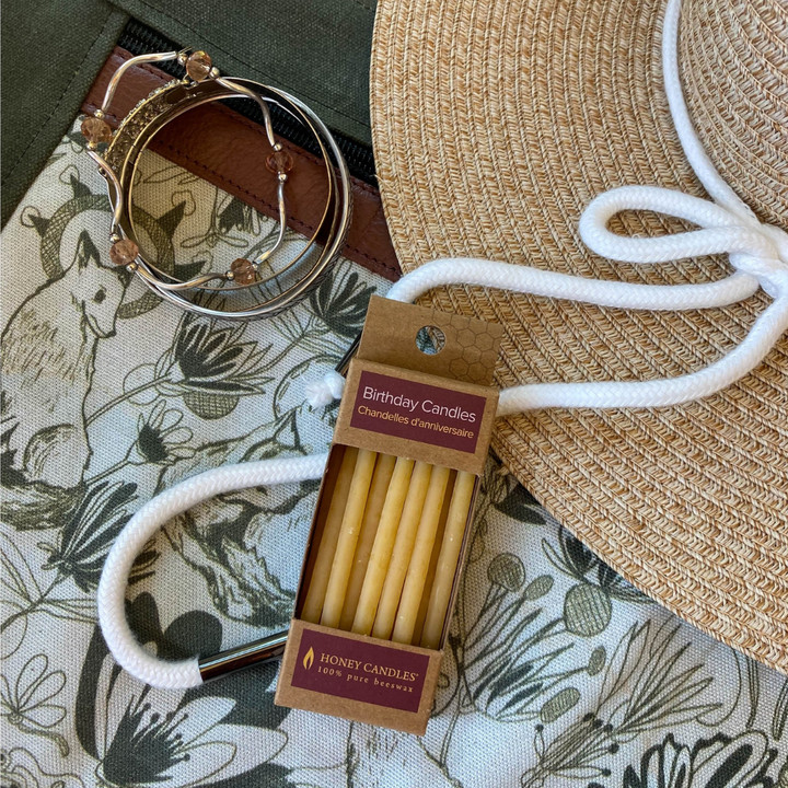 A cardboard box of birthday candles resting on a green canvas bag with a floral pattern. The box is tied to a white rope and there is a straw hat and a bracelet nearby.