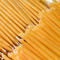 A close-up of a group of thin, yellow beeswax candles arranged tightly together. The candles have a slightly curved shape and a wick at one end.