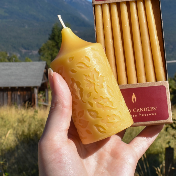 A person's hand holding a small, yellow beeswax candle with a textured leaf pattern. In the background, there is a box of beeswax candles and a scenic landscape with mountains and a cabin.