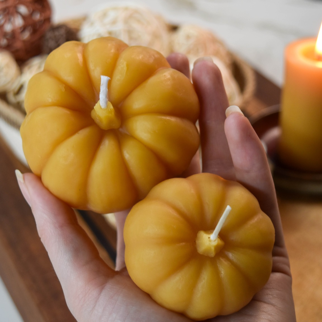 Two small, pumpkin-shaped beeswax candles in a person's hand. The candles are orange with detailed ridges and a wick in the center.