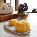 Two small, pumpkin-shaped beeswax candles on a white ceramic tray. There is a vase with flowers and a basket in the background.