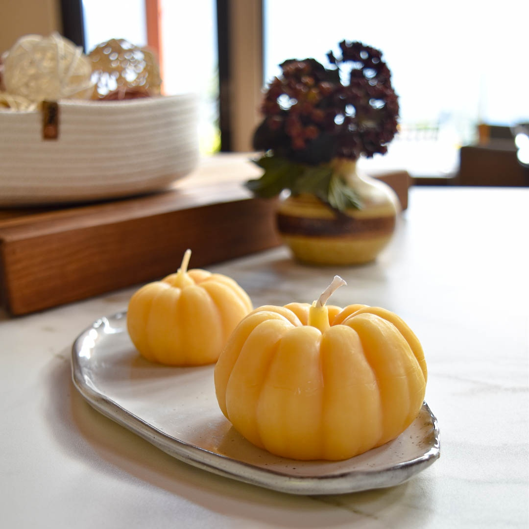 Two small, pumpkin-shaped beeswax candles on a white ceramic tray. There is a vase with flowers and a basket in the background.