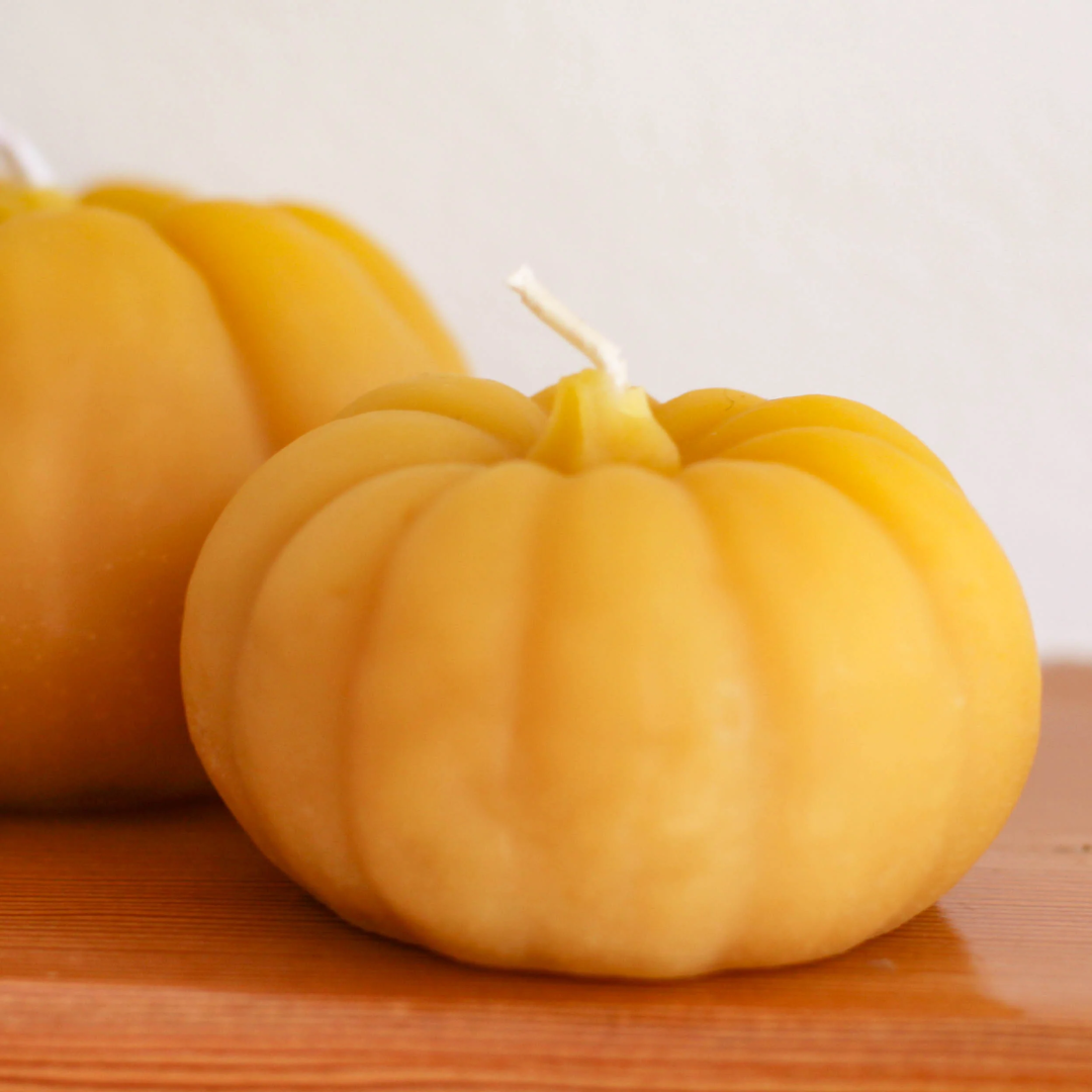 Two pumpkin-shaped beeswax candles on a wooden surface. The candles are orange with detailed ridges and a wick in the center. One candle is larger than the other.