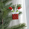 A Christmas ornament shaped like a red brick chimney with white trim and two green-and-white Santa-style legs sticking out, hanging on a green Christmas tree branch in front of a white curtain.