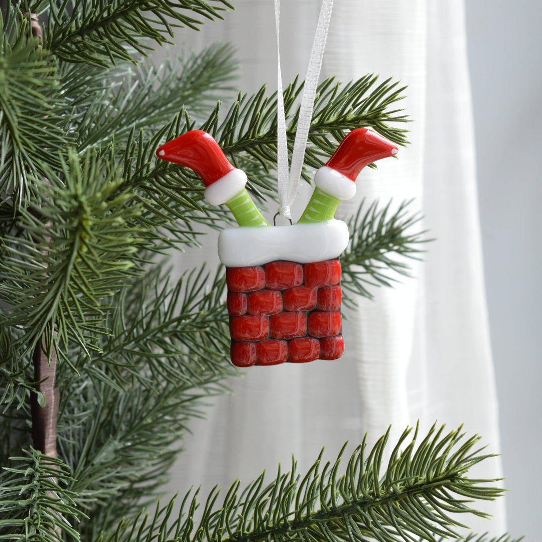 A Christmas ornament shaped like a red brick chimney with white trim and two green-and-white Santa-style legs sticking out, hanging on a green Christmas tree branch in front of a white curtain.