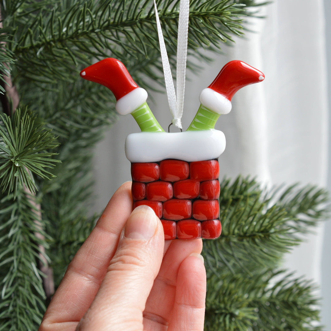 A hand holds a Christmas ornament shaped like a red brick chimney with two green and red Santa-style legs sticking out of the top, with evergreen branches in the background.