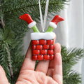 A hand holding a Christmas ornament shaped like a red brick chimney with green stockings sticking out of the top, white trim, and a white hanging ribbon. Pine branches and a light curtain are visible in the background.