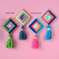 Four colorful cross-stitch ornaments displayed on a pink background. Each ornament features a diamond-shaped design with a tassel hanging below. The ornaments are labeled "Natural" and "Tropical" and showcase different color palettes, including cool blues and greens and warm oranges and pinks.