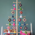 A wooden Christmas tree stand displaying colorful cross-stitch ornaments. The ornaments are diamond-shaped with tassels and feature various patterns in bright colors like pink, blue, and yellow. There are wrapped gifts and candles at the base of the tree.