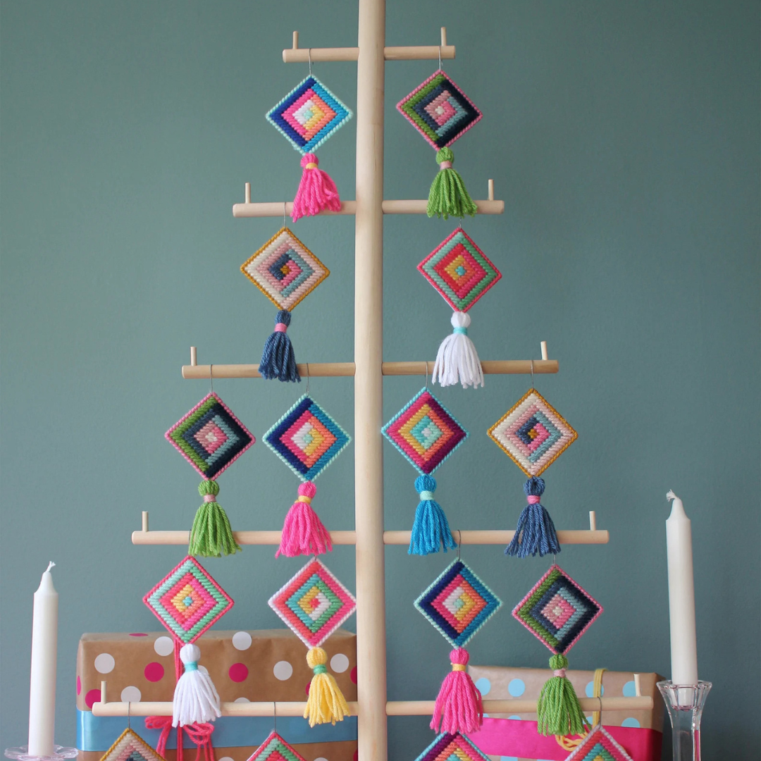 A wooden Christmas tree stand displaying colorful cross-stitch ornaments. The ornaments are diamond-shaped with tassels and feature various patterns in bright colors like pink, blue, and yellow. There are wrapped gifts and candles at the base of the tree.
