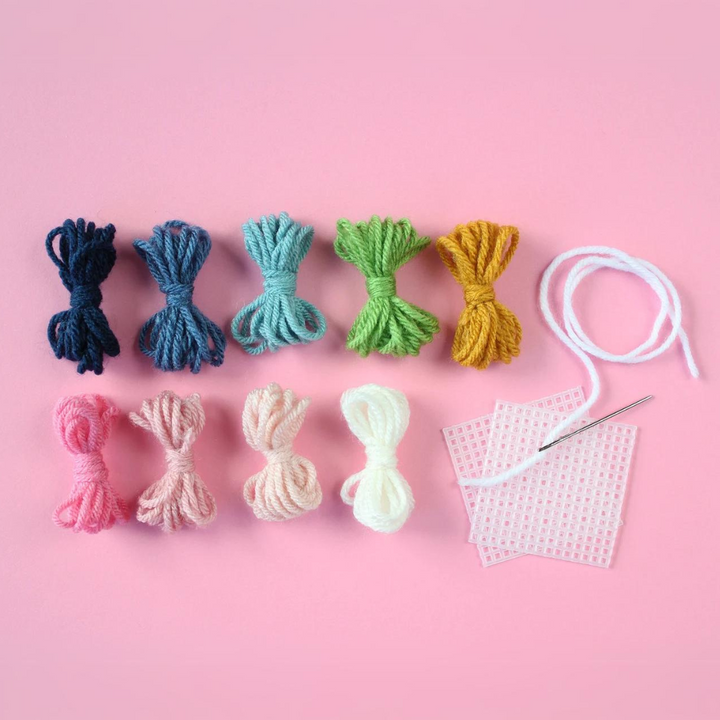 A flat lay of a DIY embroidery kit on a pink background. The kit includes colorful embroidery floss in various shades of blue, green, pink, and white. There is also a piece of white aida cloth and an embroidery needle.