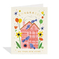 A rectangular greeting card with a cream background, featuring a whimsical illustration of a pink house surrounded by blue birds, yellow and orange flowers, balloons, and the words 'CONGRATS!' and 'ON YOUR NEW HOME'.