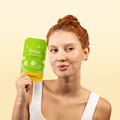 A woman with red hair in a bun holds up a green and yellow package of cryo face mask gel by waybeauty against a light beige background.