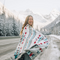 The Nibi Reversible Blanket by Mini Tipi, featuring bold Indigenous-inspired geometric patterns in teal, red, brown, and white, wrapped around a person in the mountains. This reversible and stylish blanket is available in Ottawa, Ontario, Canada.