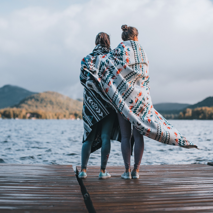The Nibi Reversible Blanket by Mini Tipi, featuring bold Indigenous-inspired geometric patterns in teal, red, brown, and white, wrapped around two people standing on a dock next to a lake. This reversible and stylish blanket is available in Ottawa, Ontario, Canada.