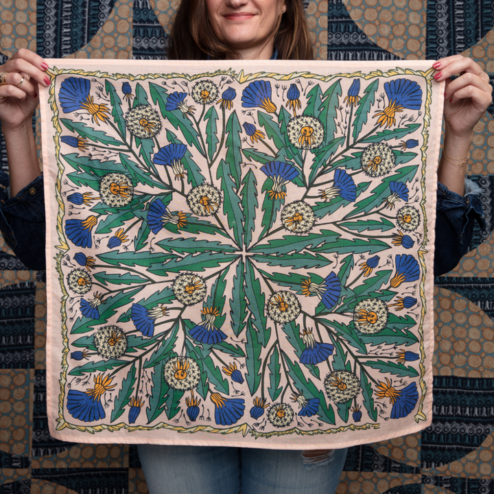 A pink bandana with a floral pattern. The pattern features blue and yellow dandelions with green leaves. The border of the bandana has a decorative, repeating pattern.