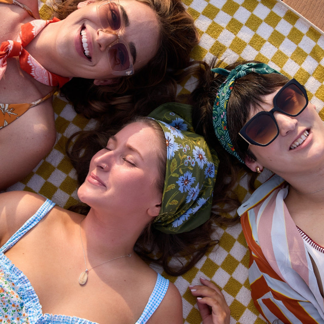 Three women lying on a checkered blanket, smiling and wearing sunglasses.