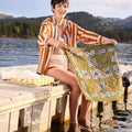 Woman holding a floral towel by a lake with mountains in the background